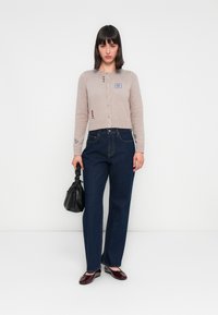 Cárdigan de punto color beige claro con botones y detalles bordados. Jeans rectos de color azul oscuro, bolso texturizado negro, zapatos burdeos.