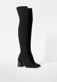 Botas negras hasta el muslo hechas de un tejido suave, con punta cuadrada y tacón grueso y brillante. Presentan un diseño sin costuras y una textura suave.