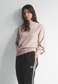 Hele roosa kõrge kaelusega kampsun koos lõdva lõike ja soonikkätistega, mida on kombineeritud mustade pükstega, millel on valged vertikaalsed triibud ja logo.