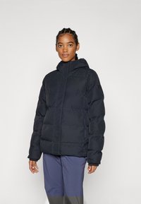 Vadderad svart jacka med dragkedja fram och huva, med en texturerad tyg och subtil branding på ärmen. Buren över mörka byxor.