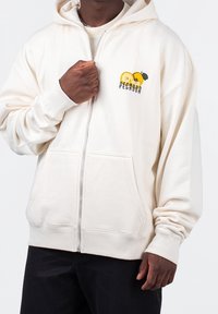 Uomo che indossa una felpa con zip color crema, con tasche frontali e una grafica di pesca gialla con il testo "Pecadora" sul lato sinistro del petto.