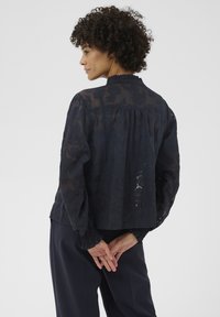 Femme aux cheveux bouclés portant un chemisier transparent à motifs floraux foncés et un pantalon ample foncé, debout avec les mains croisées dans le dos.