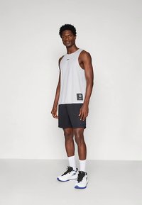 Νεαρός άνδρας στέκεται, φοράει ανοιχτό γκρι αμάνικο αθλητικό μπλουζάκι, μαύρες βερμούδες, λευκές κάλτσες και λευκά αθλητικά παπούτσια με μπλε σόλες.