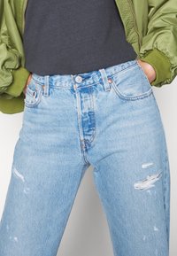 Ljusblå denimjeans med hög midja, slitna detaljer och en avslappnad passform. Matchas med en mörkgrå skjorta och en olivgrön bomberjacka.