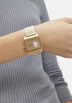 Montre numérique couleur or avec un cadran hexagonal, affichage blanc et bracelet en métal texturé, portée sur un poignet avec une manche grise côtelée.