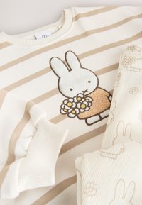 Pull rayé beige et blanc pour enfants avec un lapin tenant des fleurs brodé sur le devant, associé à des leggings crème avec des motifs lapin et fleurs.