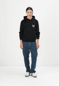 Black hooded sweatshirt with a front pocket and white logo detail. Worn with blue denim jeans and white sneakers with black accents.