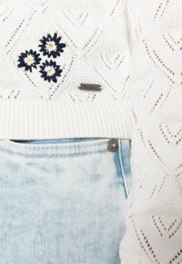Vit stickad tröja med diamantmönster, med broderade prästkragar; matchad med ljusblå urblekta jeans som har en knappdetalj.