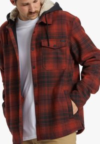 Man wearing red and black plaid button-up jacket with sherpa collar over a white shirt, looking down with one hand in pocket.
