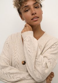 Beige strikket cardigan med V-hals, tekstureret zigzag-mønster og en stor brun knap. Blødt stof med brede ærmer.