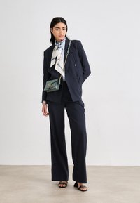 Blazer doppiopetto blu navy abbinato a pantaloni a gamba larga, insieme a una sciarpa di seta a fantasia e una piccola borsa a tracolla verde. Tacchi aperti neri.