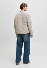 Veste matelassée beige avec un col rond, vue de dos, associée à un jean en denim bleu ample et des chaussures beiges. Texture lisse.