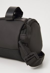Primo piano di un angolo di una borsa cilindrica nera con cerniera, tracolla regolabile e un gancio in plastica etichettato "RAINS" su sfondo bianco.