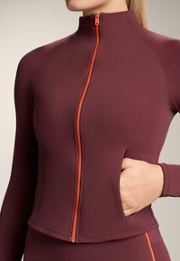 Bordó sportdzseki magas gallérral, narancssárga cipzárral és oldalzsebbel. Simább, rugalmas anyagból készült a kényelem és a mozgásszabadság érdekében.