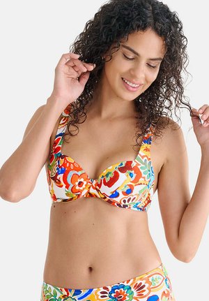 Femme aux cheveux bouclés portant un bikini floral coloré, souriante et touchant ses cheveux sur un fond clair uni.