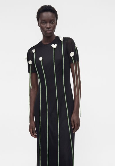 Γυναίκα φοράει μαύρο φόρεμα με διάφανα μανίκια, διακοσμημένο με κάθετα πράσινα στελέχη και μικρά λευκά λουλούδια που κρέμονται από τους ώμους και την κορμο.