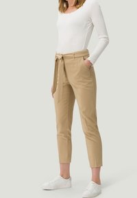Femme portant un pantalon tapered taille haute beige avec une ceinture en tissu nouée à l'avant, un haut à manches longues blanc et des baskets blanches, se tenant avec les mains dans les poches.