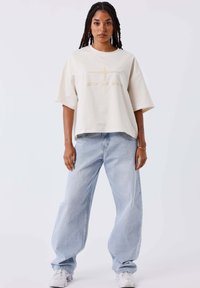 T-shirt oversize color crema con logo ricamato, abbinata a jeans baggy azzurri e scarpe da ginnastica bianche. Tessuto morbido, vestibilità rilassata.
