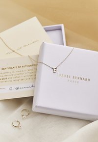 Goldene Halskette mit einem Anhänger in Form des Buchstabens "B", präsentiert auf einem weichen Stoffhintergrund zusammen mit einem Zertifikat und einer weißen Schachtel mit goldener Schrift.