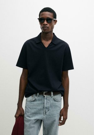 Muž v černém polo tričku s krátkým rukávem, světlých modrých džínách, tmavých slunečních brýlích, držící složenou vínově červenou bundu na jednobarevném bílém pozadí.