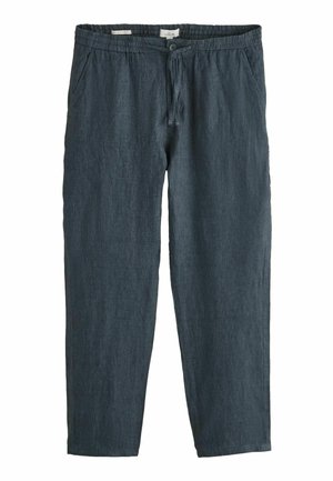 Sötétkék lenvászon nadrágot viselő férfi, gombos hátsó zsebbel és egy fehér, hosszú ujjú inget betűrve.