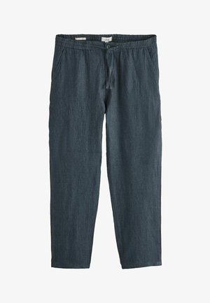 Sötétkék lenvászon nadrágot viselő férfi, gombos hátsó zsebbel és egy fehér, hosszú ujjú inget betűrve.