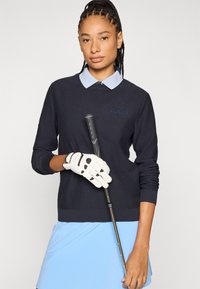 Sötétkék kötött golfpulóver világoskék gallérral, logóval díszítve. A modell egy golfütőt tart, fehér kesztyűt viselve, fogantyú részletekkel.