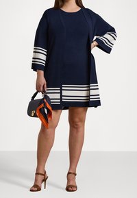 Merenkukkaa oleva mekko, jossa on valkoisia vaakaraitoja, kyynärpituuden hihat ja rento leikkaus. Asusteena pieni käsilaukku, jossa on kuvioitu huivi.