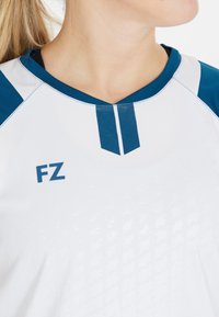 Nära bild av en person som bär en vit sporttröja med blå krage och axelaccenter, med "FZ" tryckt på bröstet.