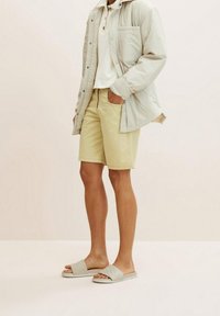 Tenue décontractée avec une doudoune grise clair, un polo blanc, un short jaune clair et des sandales à glissière grises matelassées.