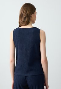 Femme aux longs cheveux bruns portant un haut sans manches en textile bleu marine et un pantalon assorti, vue de dos sur un fond uni.
