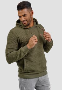 Sudadera texturizada color verde oliva con capucha, bolsillo frontal y cordones. Presenta un patrón acolchado y puños acanalados. Llevada con jeans claros desgastados.