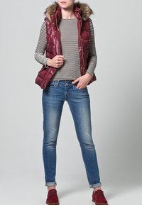 Gilet imbottito color borgogna con cappuccio rifinito in pelliccia indossato sopra una maglia a maniche lunghe a righe bianche e nere, abbinato a jeans blu e scarpe rosse.