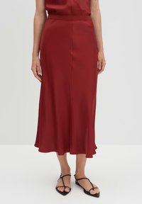 Roter Satin-Mid Rock mit glatter Textur, ausgestattet mit einem taillierten Bund und einem ausgestellten Saum. Kombiniert mit schwarzen, flachen Sandalen mit Riemen.