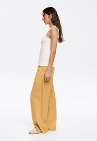 White sleeveless tank top, paired with light yellow wide-leg linen pants, featuring a pocket and frayed hems. Neutral sneakers complete the look.