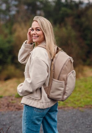 Beiger Rucksack mit mehreren Reißverschlussfächern, gepolsterten Trägern und strukturiertem Material. Getragen über einem leichten Pullover und Jeans. Grüner Hintergrund.