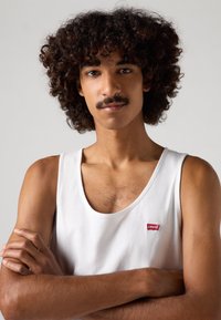 Fehér pamut ujjatlan póló, kerek nyakkivágással, bal oldalon egy kis piros Levi's logóval. A modellen göndör haj van, karjai keresztbe állnak.