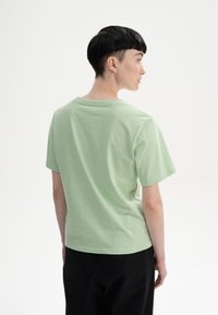 Helles grünes Baumwoll-T-Shirt, kurze Ärmel, runder Halsausschnitt, lockere Passform, sichtbare Nähte am Saum, einfarbige Textur, getragen mit schwarzen Hosen.