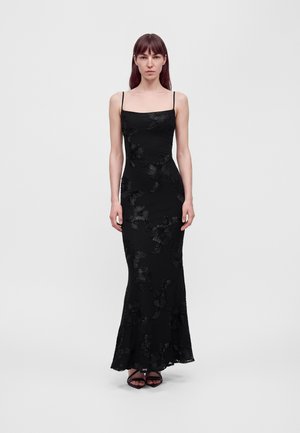 Une femme présente une robe noire à fines bretelles, longue jusqu’au sol et ornée de motifs floraux, sur un fond uni et clair.