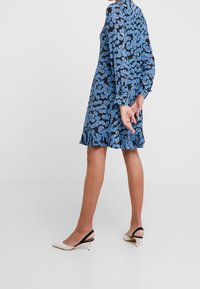 Sininen kukallinen mekko pitkillä hihoilla ja röyhelöhelmallahan, jota komistaa musta tausta. Mallilla on jalassaan kermaiset korokepohja-nilkkurit mustilla hihnoilla.