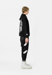 Sudadera negra con diseño de plumas plateadas en la parte trasera, combinada con pantalones negros que presentan detalles de plumas blancas. El modelo lleva botas de combate blancas.