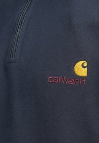 Μπλε ναυτικό φλις μπλουζάκι με  φερμουάρ μέχρι το στήθος και ριμπ υφή. Φέρει κίτρινο λογότυπο σε σχήμα στροβίλου και το "carhartt" κεντημένο σε μπορντό.