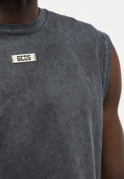 Αμάνικο σκουρόχρωμο γκρι μπλουζάκι με ελαφρώς ξεβαμμένη όψη και μια μικρή λευκή ετικέτα με την επιγραφή "GCDS" στο πάνω μέρος του στήθους, φορεμένο από άτομο με σκούρο χρώμα δέρματος.