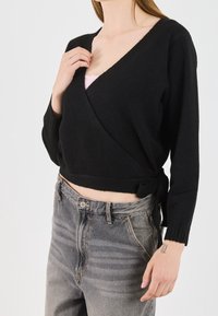 Czarny sweter typu wrap z głębokim dekoltem w kształcie V, długimi rękawami i detalem w postaci kokardy. Połączony z wyblakłymi, szarymi dżinsami o wysokim stanie. Miękka, teksturowana tkanina.