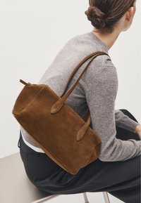 Sac à main en daim marron avec deux poignées allongées. Forme rectangulaire, texture douce et bords arrondis. Porté à l'épaule, associé à un pull gris.
