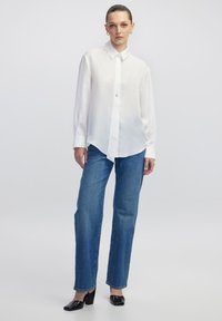 Biała koszula z długim rękawem, połączona z niebieskimi jeansami prostymi i czarnymi butami na słupku. Prosta, gładka tekstura materiału.