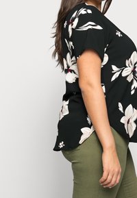 Svart blommig blus med korta ärmar, prydlig med stora vita och rosa blommor, sidofacker och en avslappnad passform, matchad med olivgröna byxor.