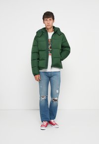 Doudoune verte avec fermeture éclair à l'avant, capuche et grandes sections matelassées ; associée à un jean bleu déchiré et des baskets rouges.