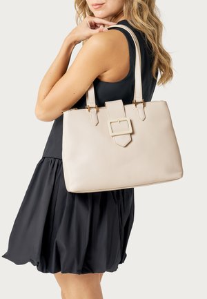 Donna con un abito nero senza maniche che porta una grande borsa beige con doppi manici e dettaglio a fibbia frontale sulla spalla.