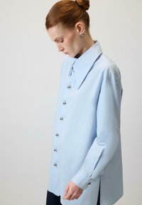 Gaiši zila krekla krekliņa ar lielu apkakli, sudraba pogām un gariem piedurknēm. Raksturīgs taisns griezums un sānu šķēlumi kustībai.